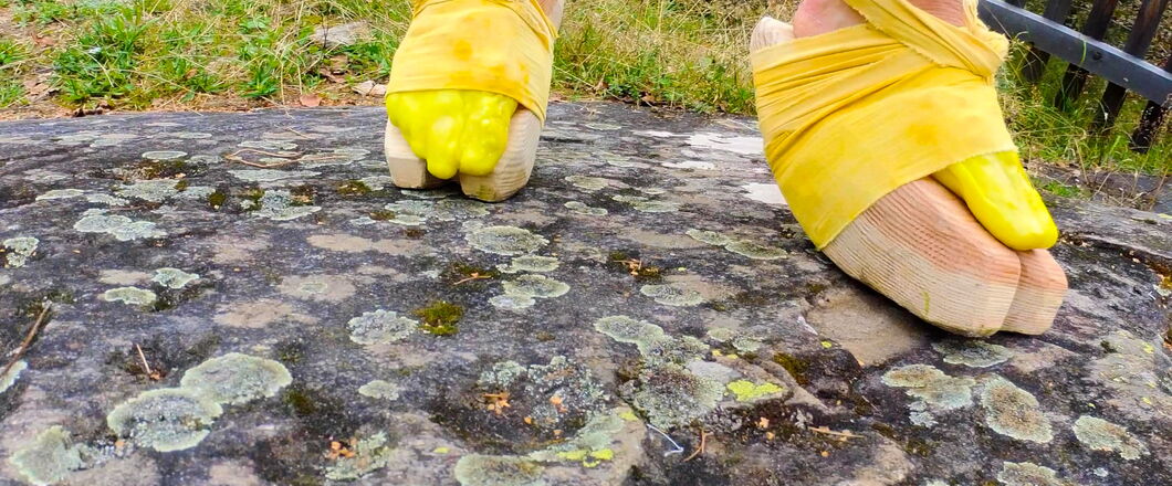 Zwei Füße sind gelb angemalt, mit gelben Stoffbändern an Holzklötzen befestigt und gehen über einen großen Stein. Link zum Profil von Eva Susova.