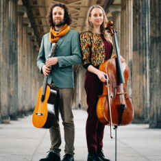 The two musicians of the duo Shiluv stand in a portico with their instruments in hand - a guitar and a cello.