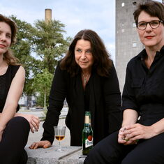 The three authors Peggy Mädler, Annett Gröschner, and Wenke Seemann are sitting on a concrete block in front of a Plattenbau building, with a wine glass and a bottle of beer in front of them.