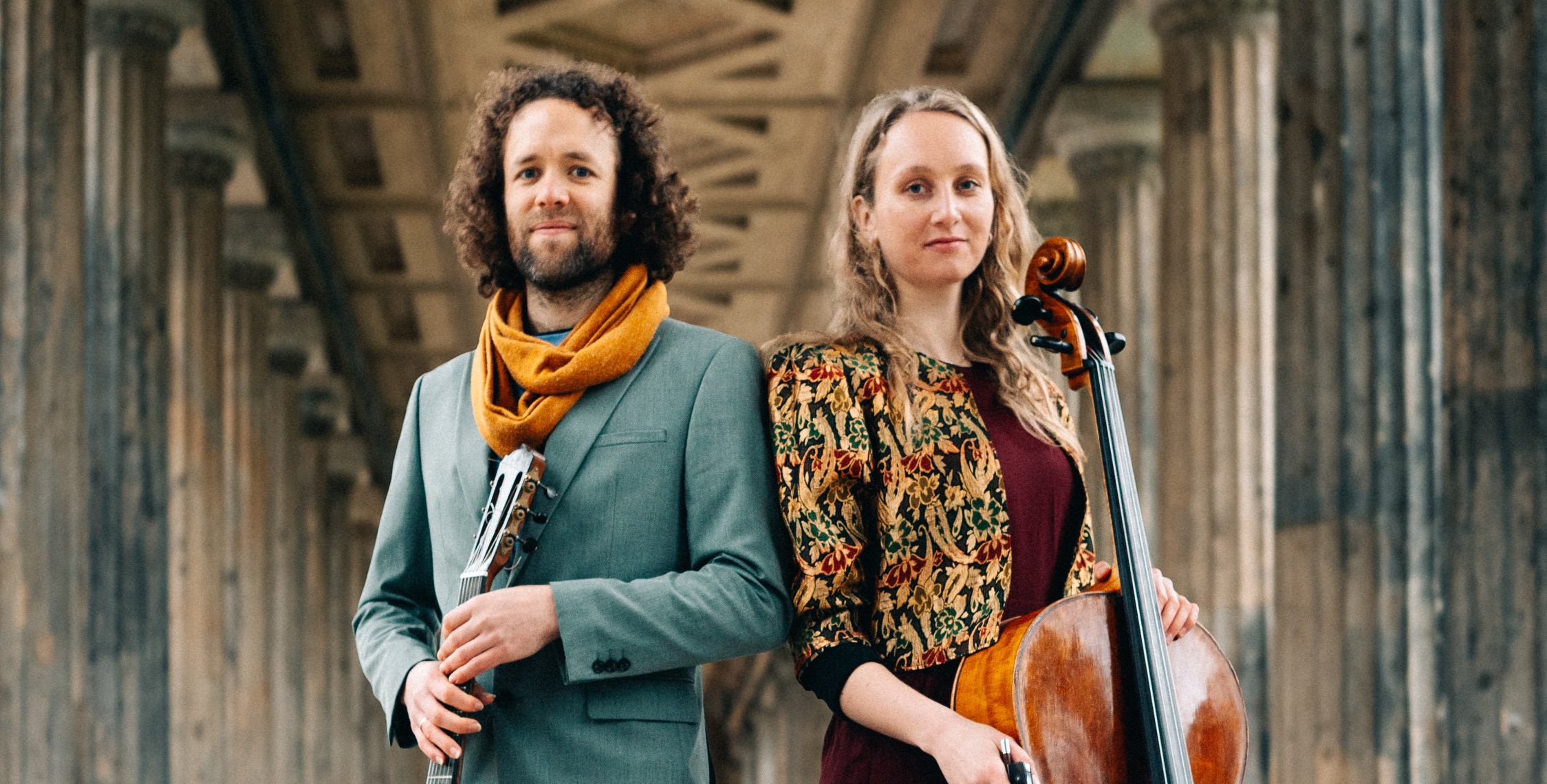 Die beiden Musiker:innen des Duo Shiluv stehen in einem Säulengang mit ihren Instrumenten in der Hand - einer Gitarre und einem Cello.