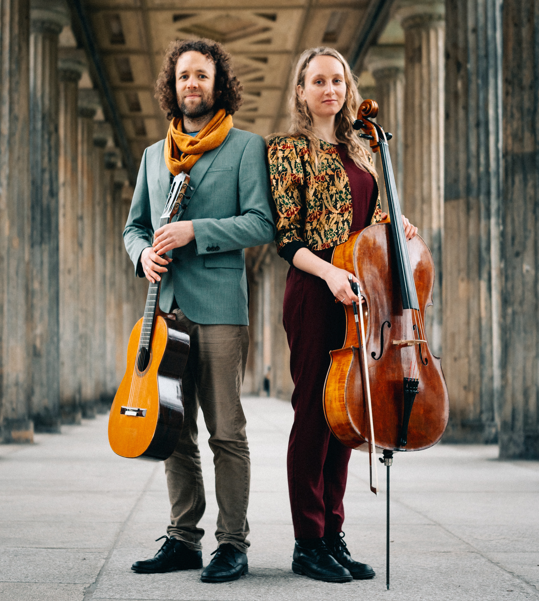 Die beiden Musiker:innen des Duo Shiluv stehen in einem Säulengang mit ihren Instrumenten in der Hand - einer Gitarre und einem Cello.
