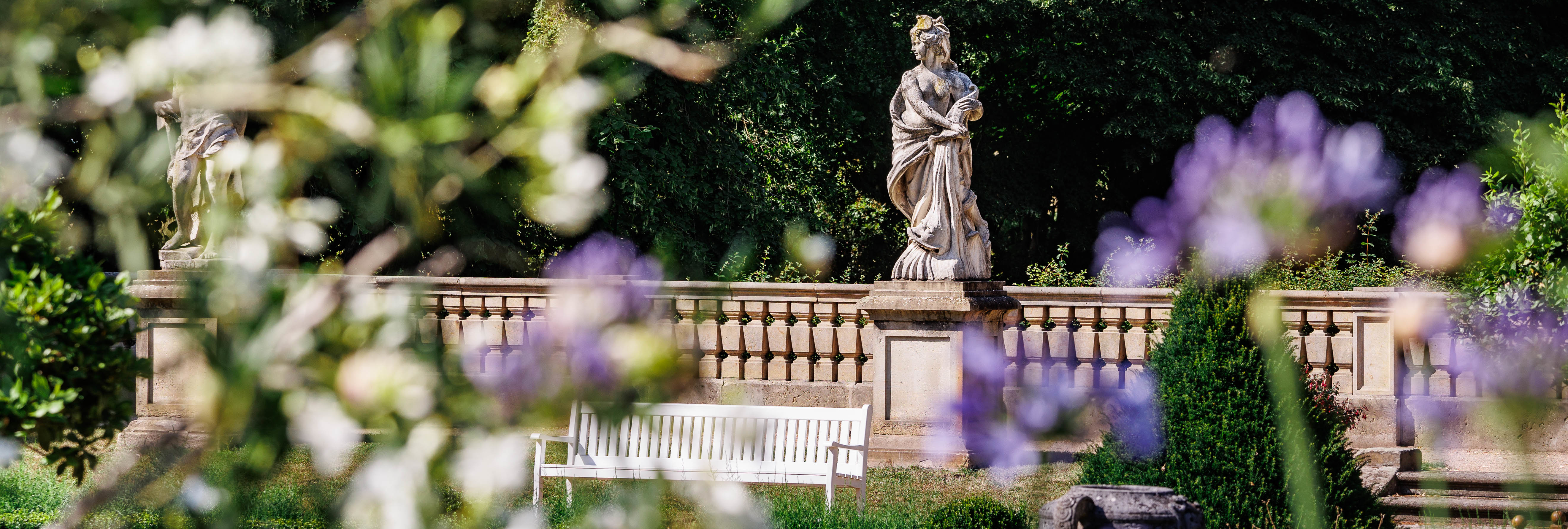 Ansicht des Parks von Schloss Wiepersdorf mit einer Skulptur im Hintergrund und einer weißen Parkbank im Vordergrund.