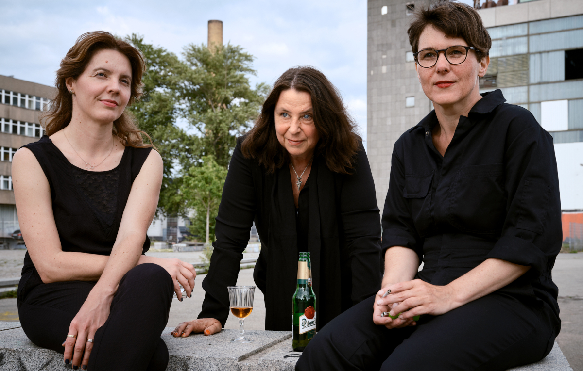Die drei Autorinnen Peggy Mädler, Annett Gröschner und Wenke Seemann sitzen auf einem Betonklotz vor einem Plattenbau, vor ihnen ein Weinglas und ein Flaschenbier
